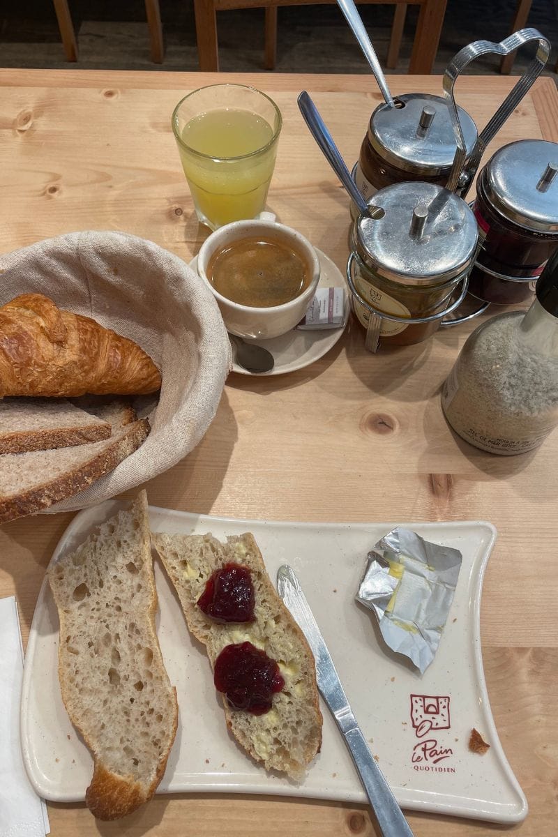 A baguette cut in two slathered with jam and butter, a basket of bread slices and croissant, a black cofee, an apple juice, and a selection of spreads